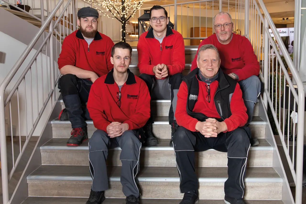 Fünf Mitarbeitende von Möbel Rommel sitzen auf einer Treppe im Möbelhaus und tragen Arbeitskleidung. Das Team steht für professionellen Lieferservice, Möbelmontage und Küchenmontage in der Region.